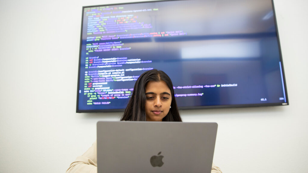 Sameera Shah types on a laptop in front of a monitor displaying lines of code.
