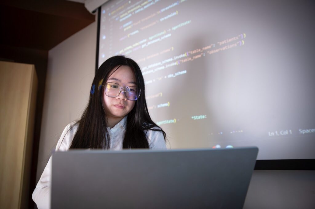Catlynh Nguyen works on a laptop while lines of code are projected on a screen behind her