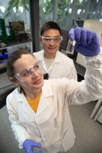 Lia Ryan examines a small vial while faculty mentor Xin Xu stands behind her in a laboratory